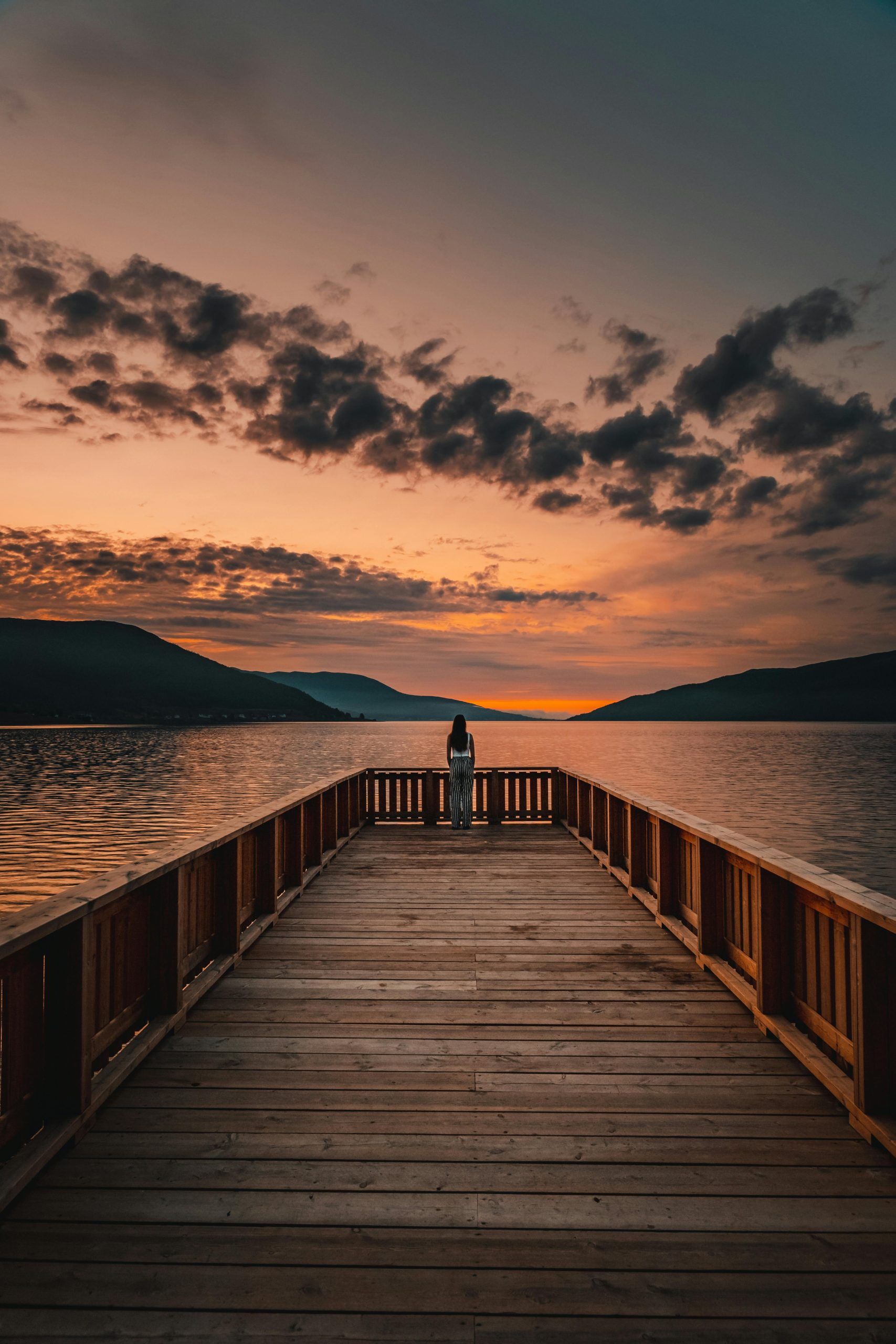A tranquil sunset view from a wooden pier overlooking a calm lake and distant mountains.